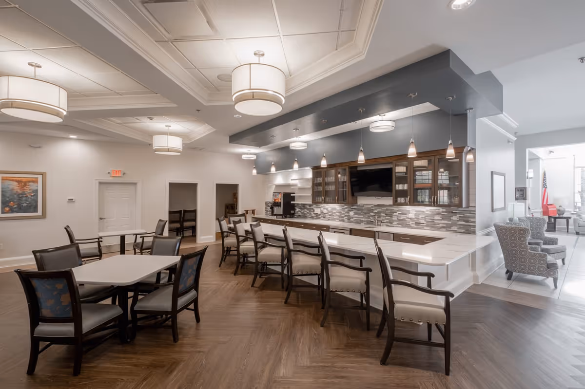 A spacious and modern dining area in a senior living facility featuring multiple tables with chairs, a long counter with high chairs, pendant and ceiling lights, and a kitchen area with cabinets and a tiled backsplash. There are also two patterned armchairs near a window with an American flag visible in the background.