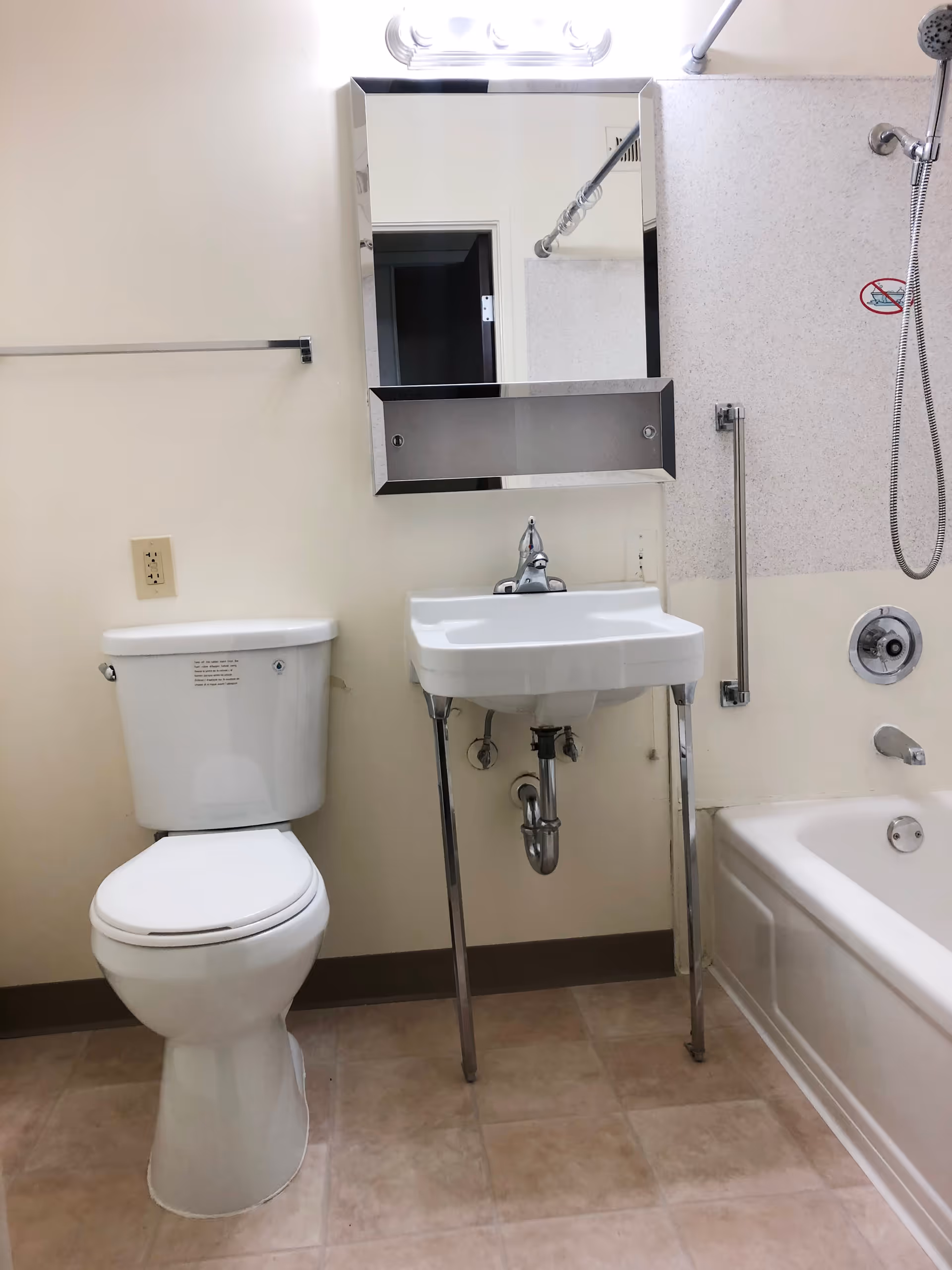 A clean bathroom featuring a white toilet, a wall-mounted sink with exposed plumbing and metal legs, a rectangular mirror above the sink, and a bathtub with a showerhead and grab bar. The walls are light-colored, and the floor has beige tiles.