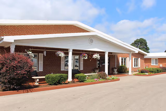 Brick single-story senior living facility entrance with white columns, hanging flower baskets, and landscaped beds.