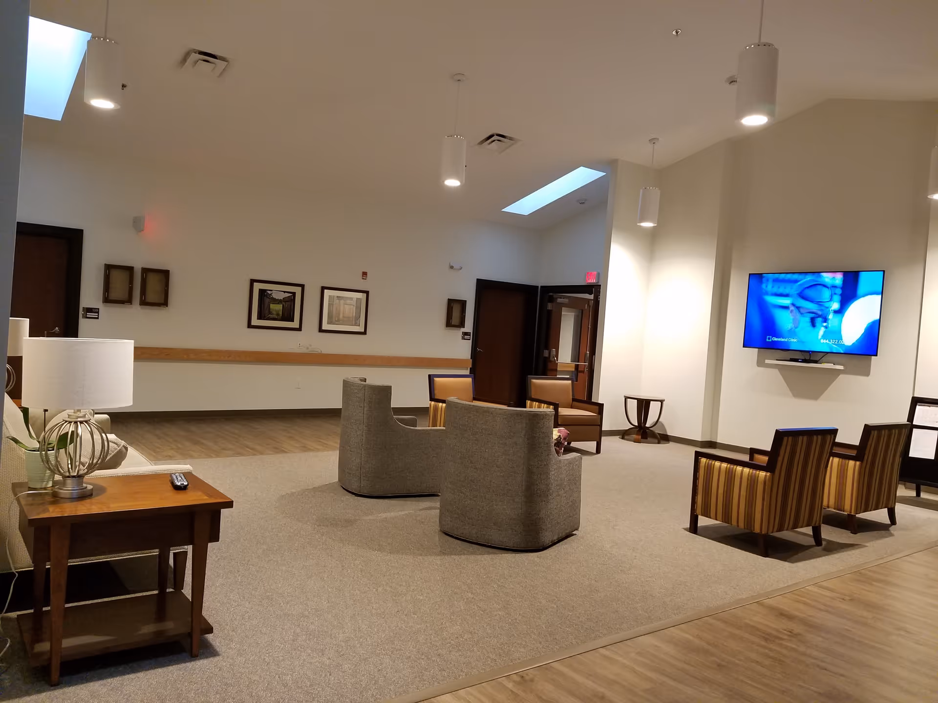 Spacious senior living lounge with seating arrangements, a wall-mounted TV, side tables and overhead lighting.