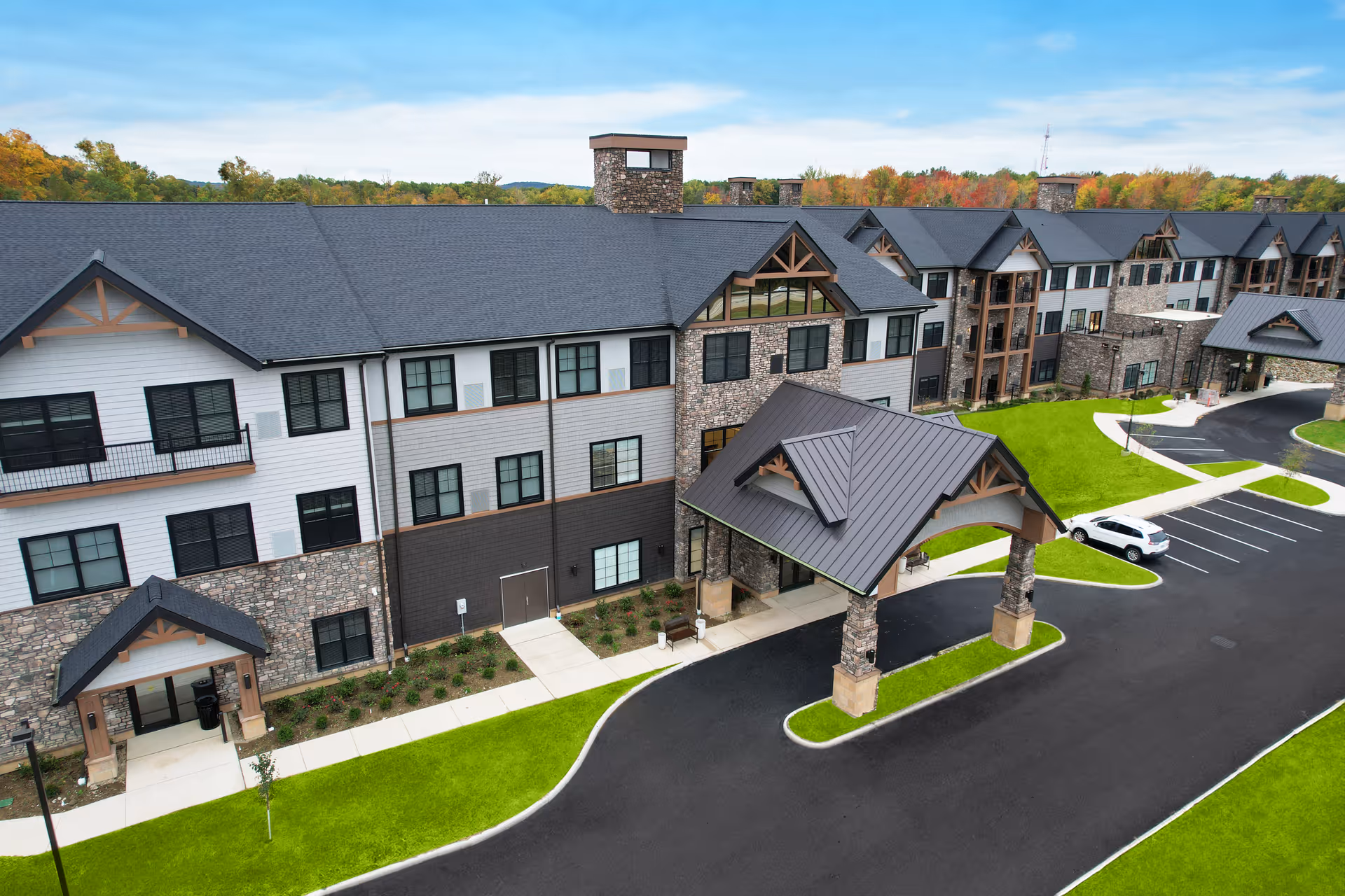 Aerial view of a large three-story senior living building with stone and siding exterior, a covered porte-cochere, parking lot and landscaped lawns.