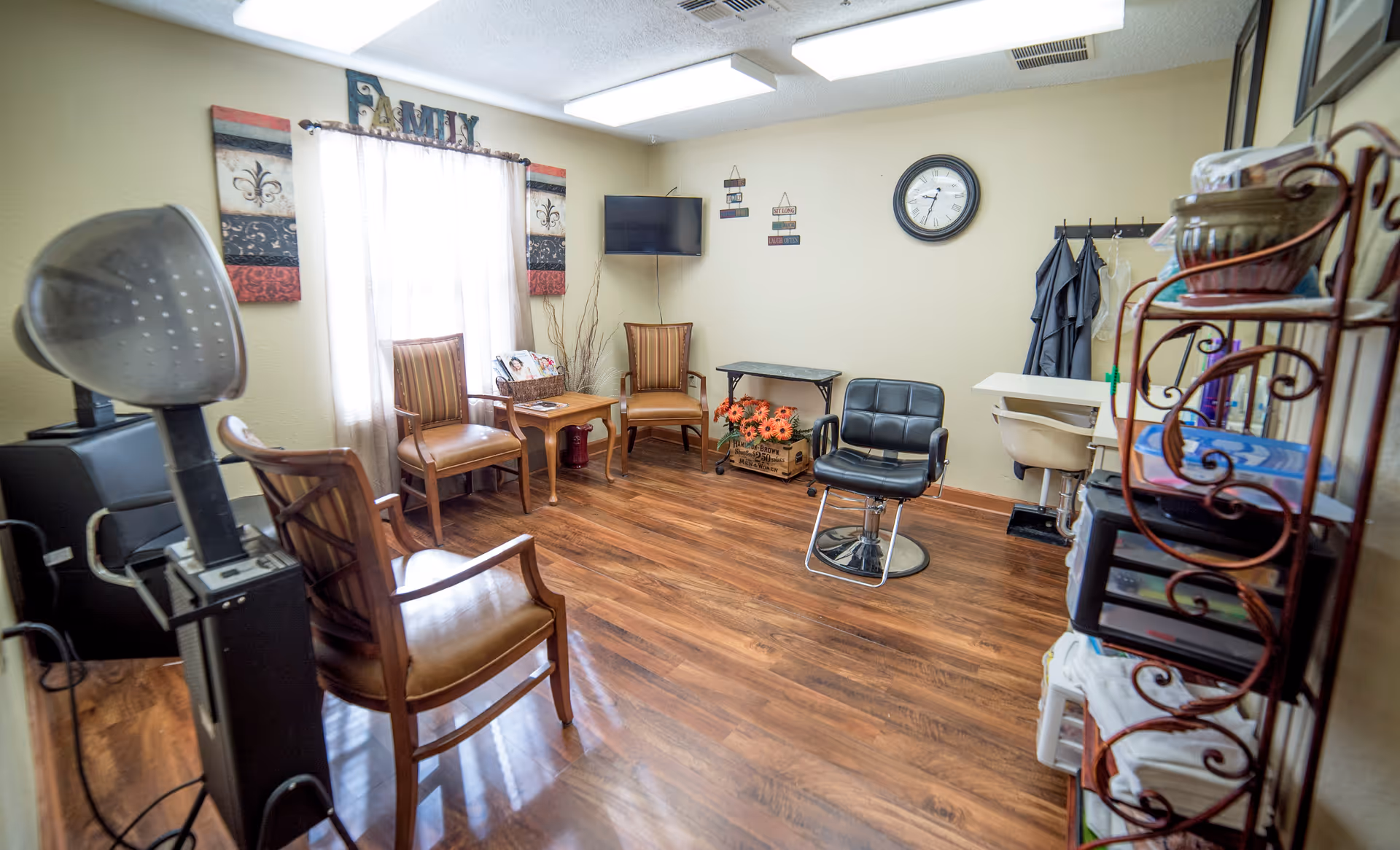 A cozy room with wooden flooring and beige walls, featuring several chairs arranged around the space including a black salon chair. There is a window with sheer white curtains and decorative wall art above it that says 'FAMILY'. A small table with flowers and a TV mounted on the wall are also visible. On the right side, there is a metal shelving unit with towels and other items, and a sink with hanging aprons or gowns nearby.