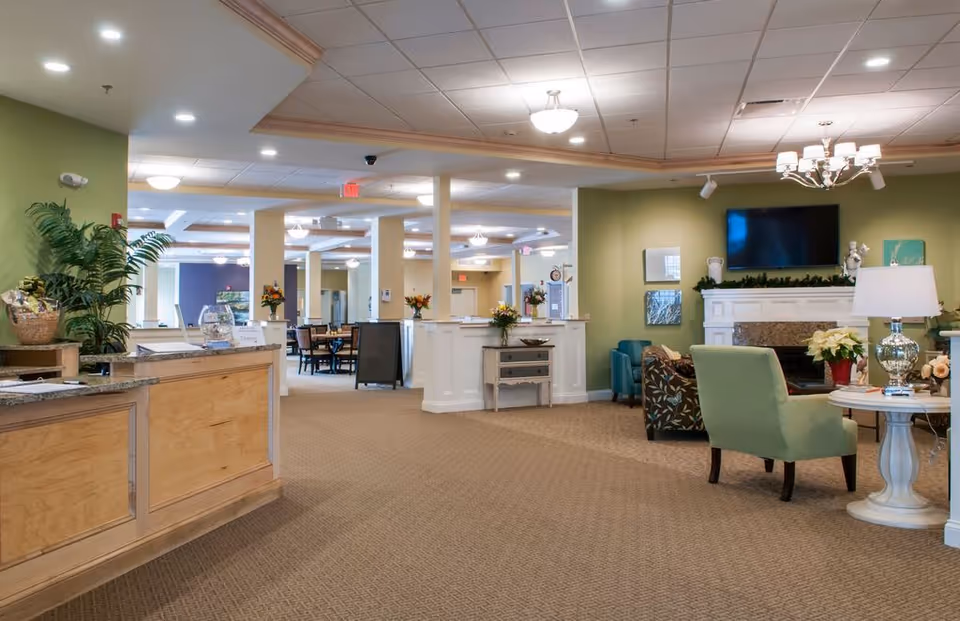 Spacious, well-lit senior living facility lobby with a reception desk, seating area with chairs and a fireplace, and a dining area visible in the background.