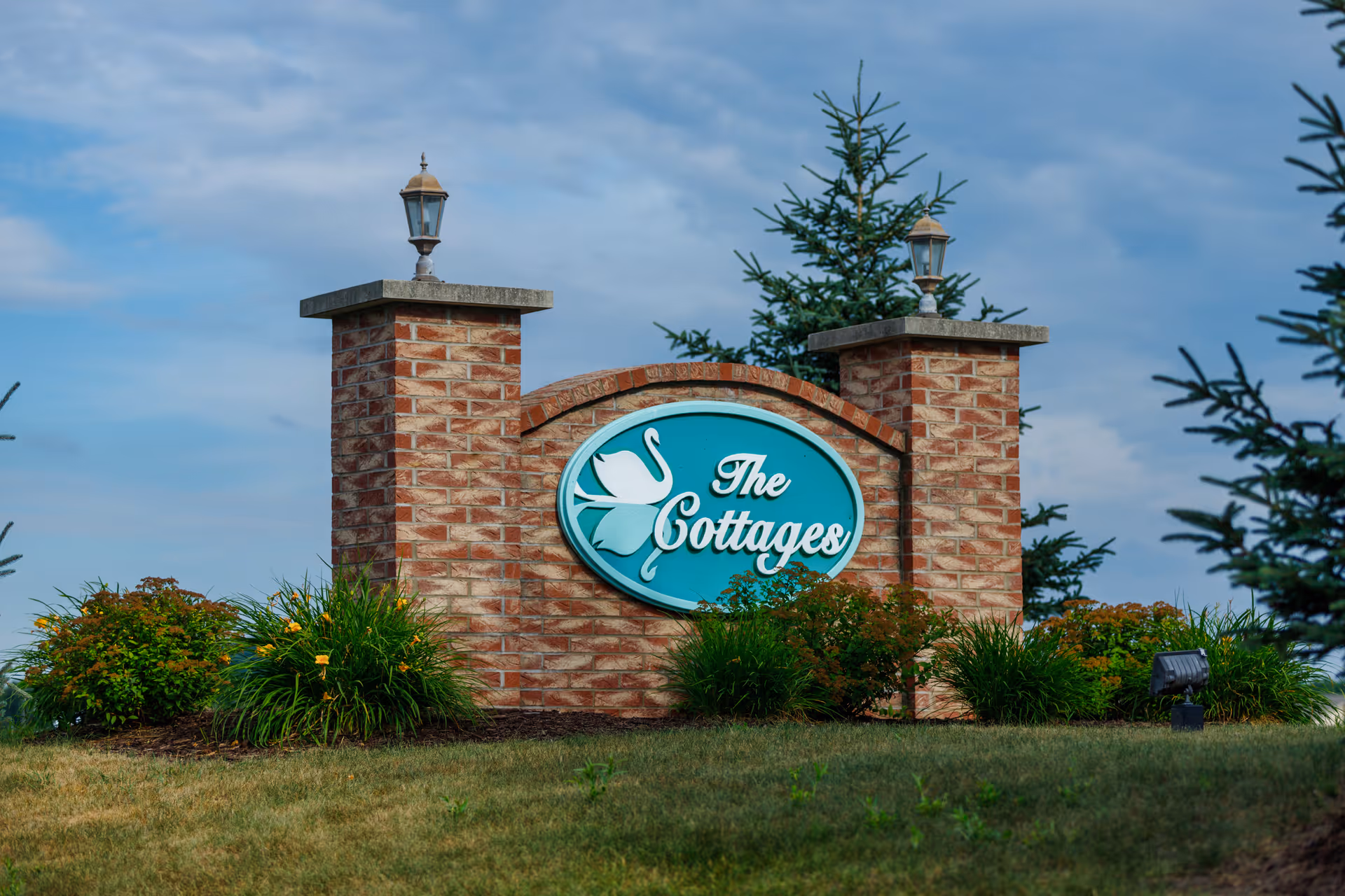 A brick sign with two pillars topped with lanterns displaying the name 'The Cottages' with a swan logo, surrounded by green shrubs and grass under a partly cloudy sky.