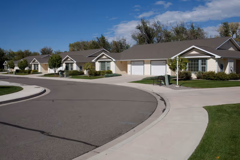 A row of single-story residential buildings with garages, surrounded by well-maintained lawns and trees under a partly cloudy blue sky.