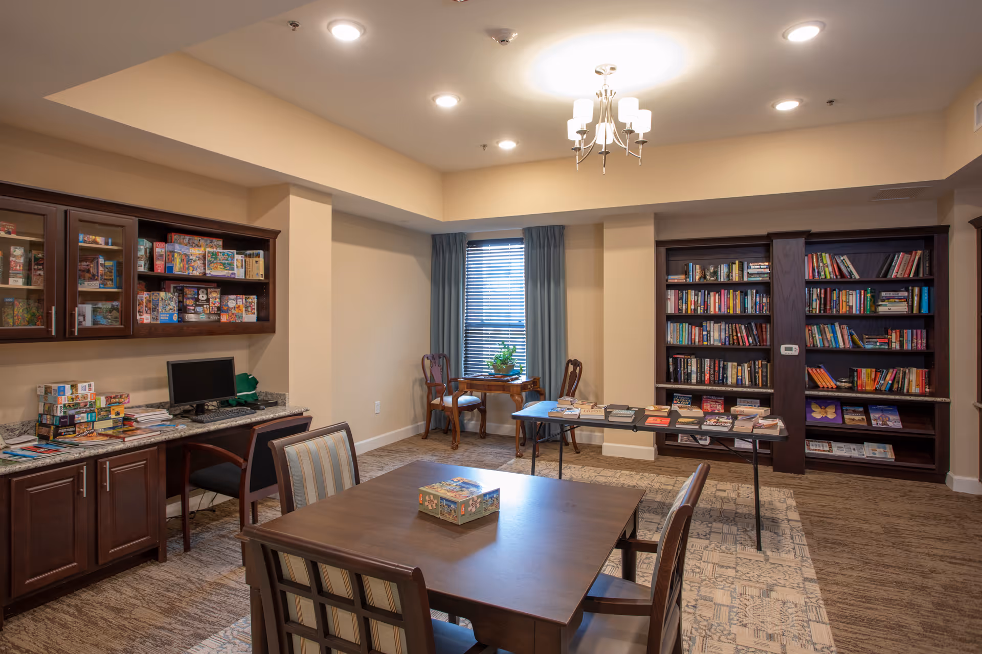 A well-lit activity room with a wooden table and chairs in the foreground, a table with books and games in the middle, bookshelves filled with books along the back wall, a window with blue curtains, and a cabinet with board games and a computer on the left side.