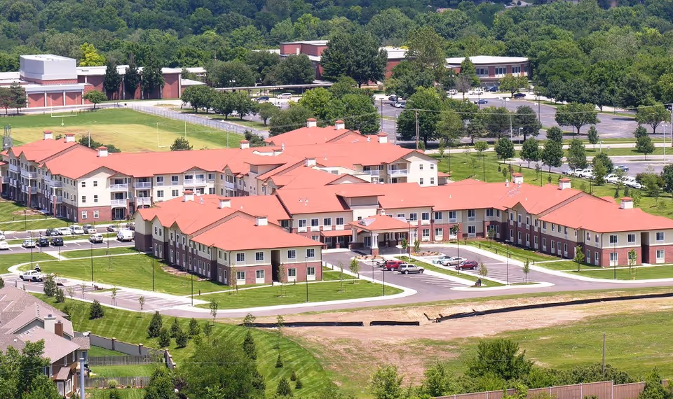 Aerial view of a large senior living facility with multiple connected buildings featuring red roofs and beige walls, surrounded by green lawns, trees, parking lots, and nearby residential houses.