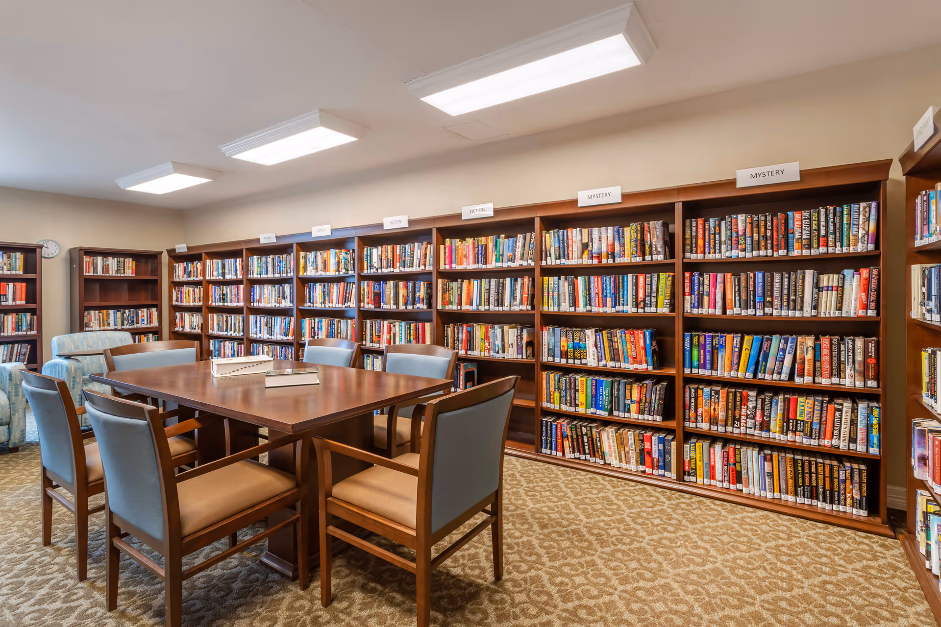 A well-lit library room with wooden bookshelves filled with books organized by categories such as fiction and mystery. In the center, there is a wooden table surrounded by six cushioned chairs. The room has a beige carpet with a patterned design and soft overhead lighting.