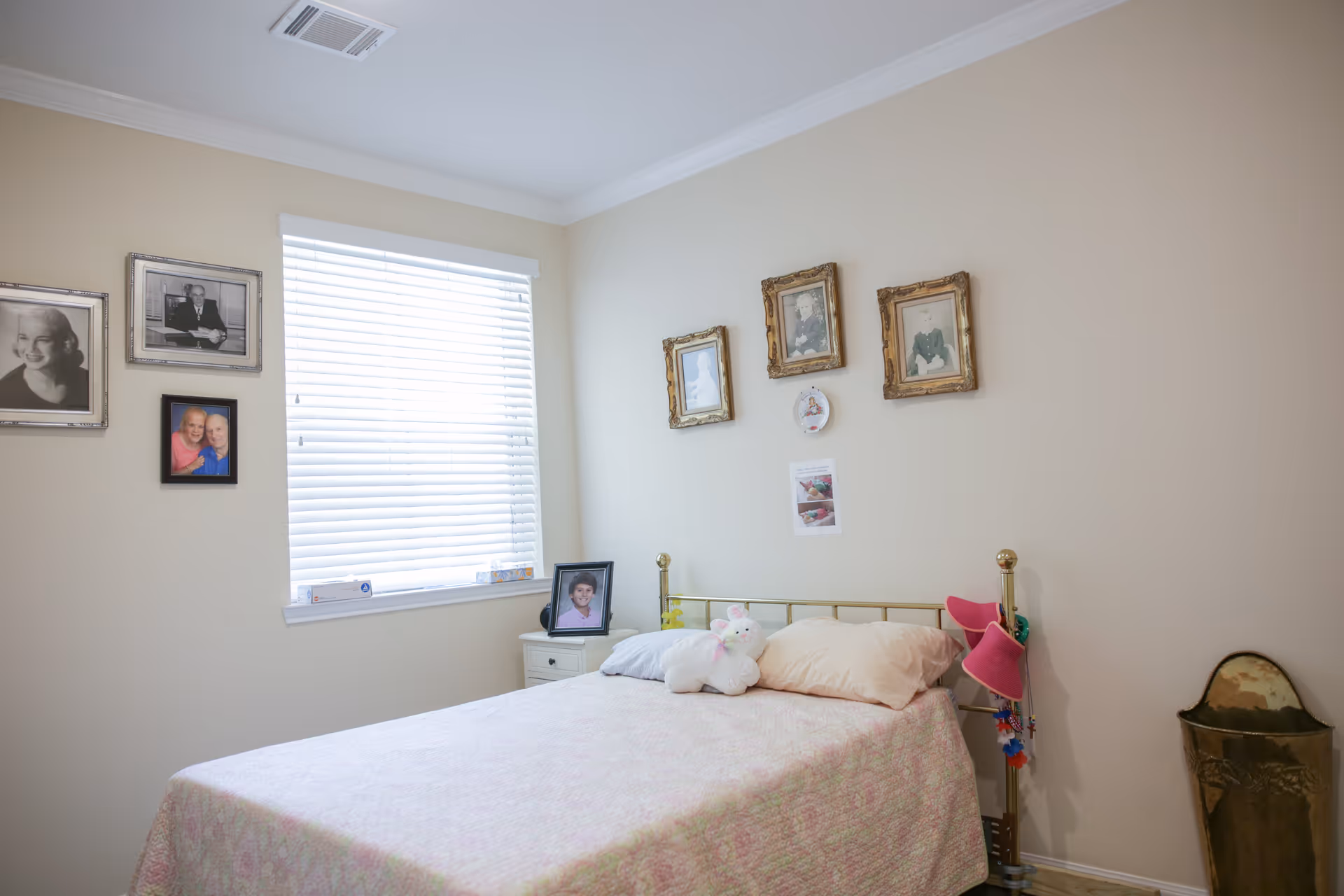 A simple single bed in a softly lit bedroom with family photos on the walls and a nightstand by a window with blinds.