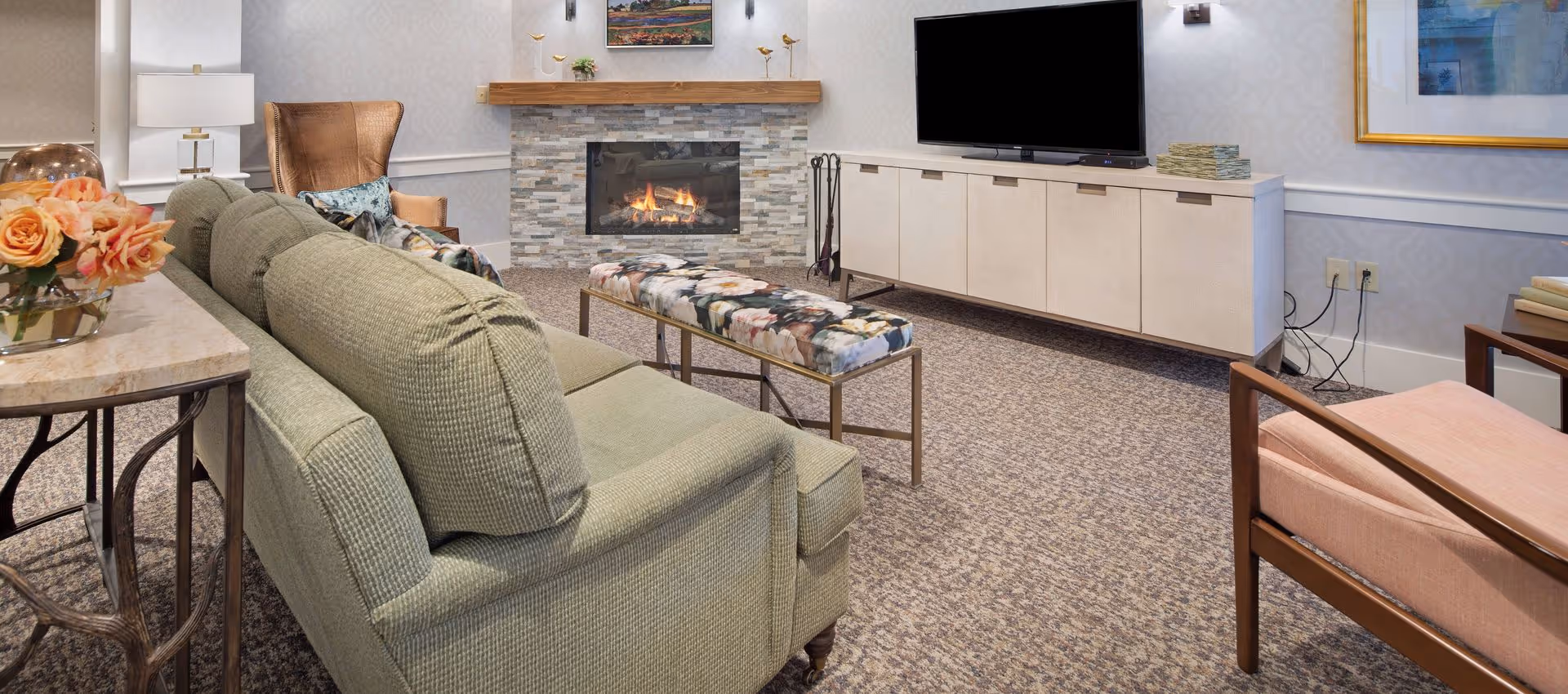 Cozy senior living common room with a green sofa facing a fireplace and wall-mounted TV, flanked by chairs and a patterned bench.