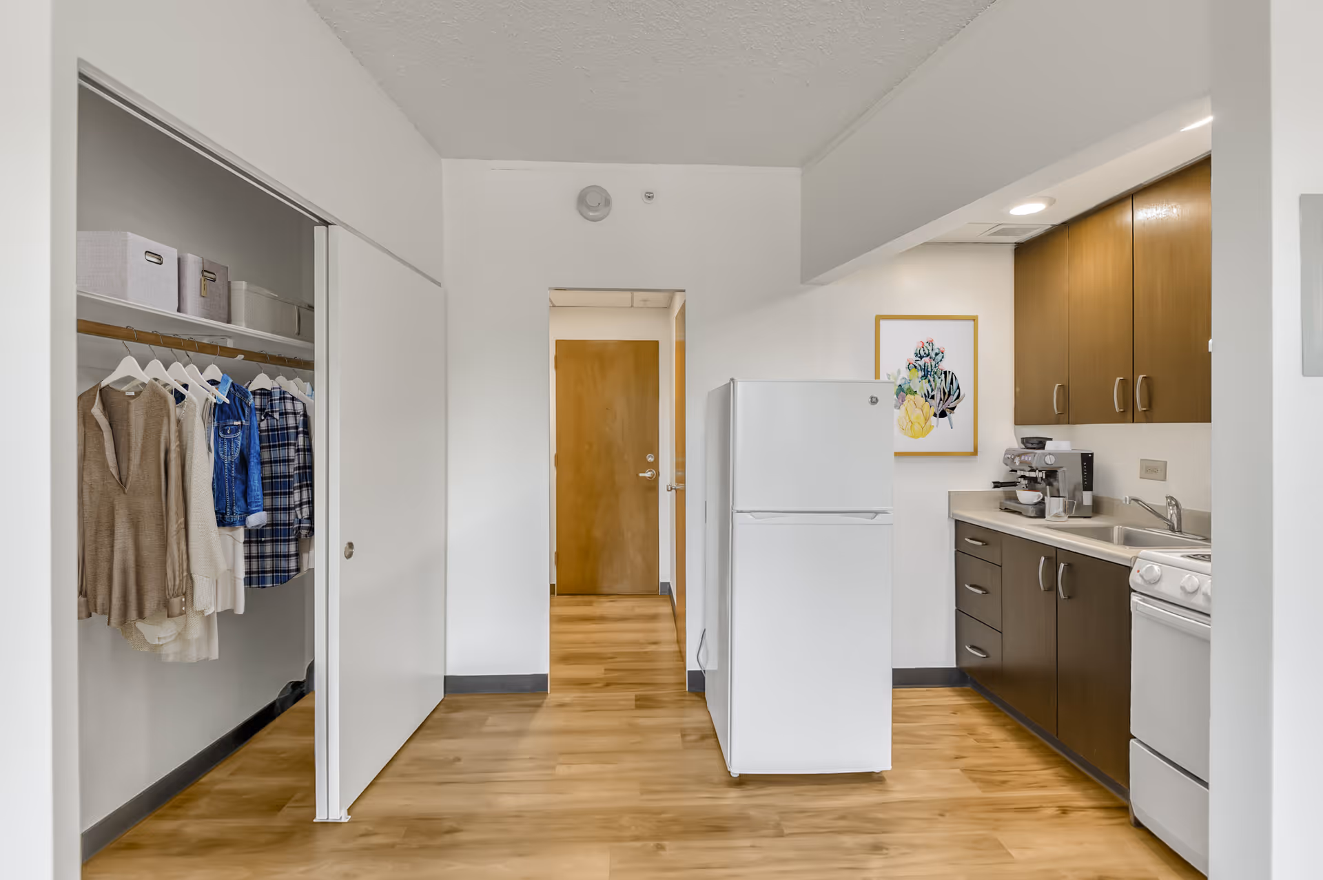 Studio apartment interior showing a kitchenette with a white refrigerator and brown cabinets, an open closet with hanging clothes, and a wooden entrance door.