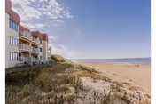 View of a sandy beach with grassy dunes in the foreground and a multi-story building with balconies on the left side under a partly cloudy blue sky.