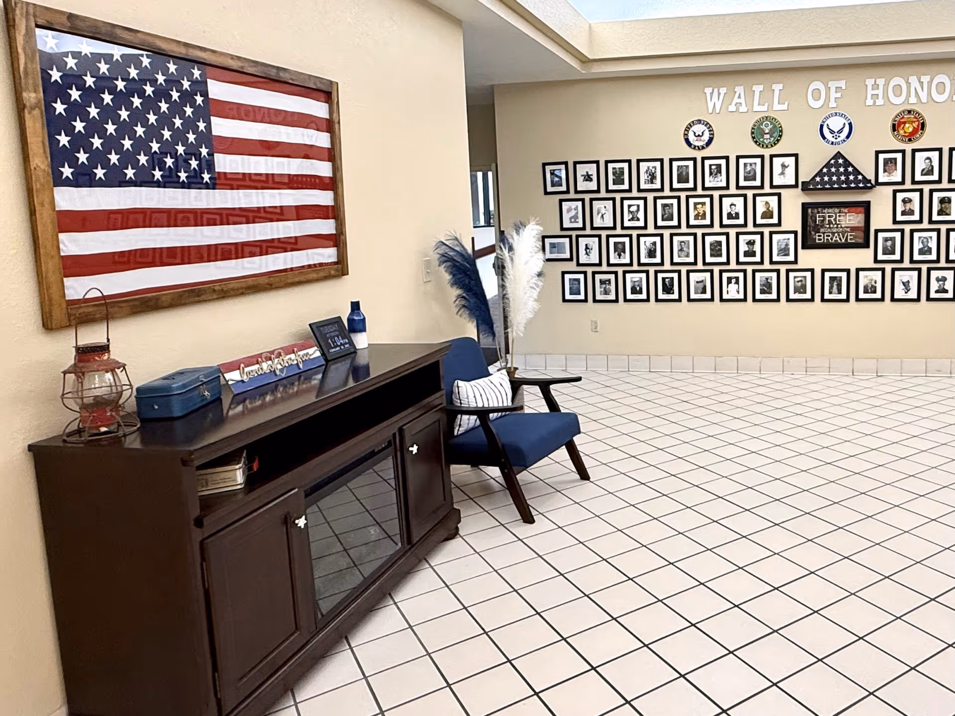 Interior view of a hallway or common area with a large framed American flag on the wall, a wooden cabinet with decorative items on top, a blue chair with a pillow, and a 'Wall of Honor' display featuring framed photos and military emblems on the opposite wall.
