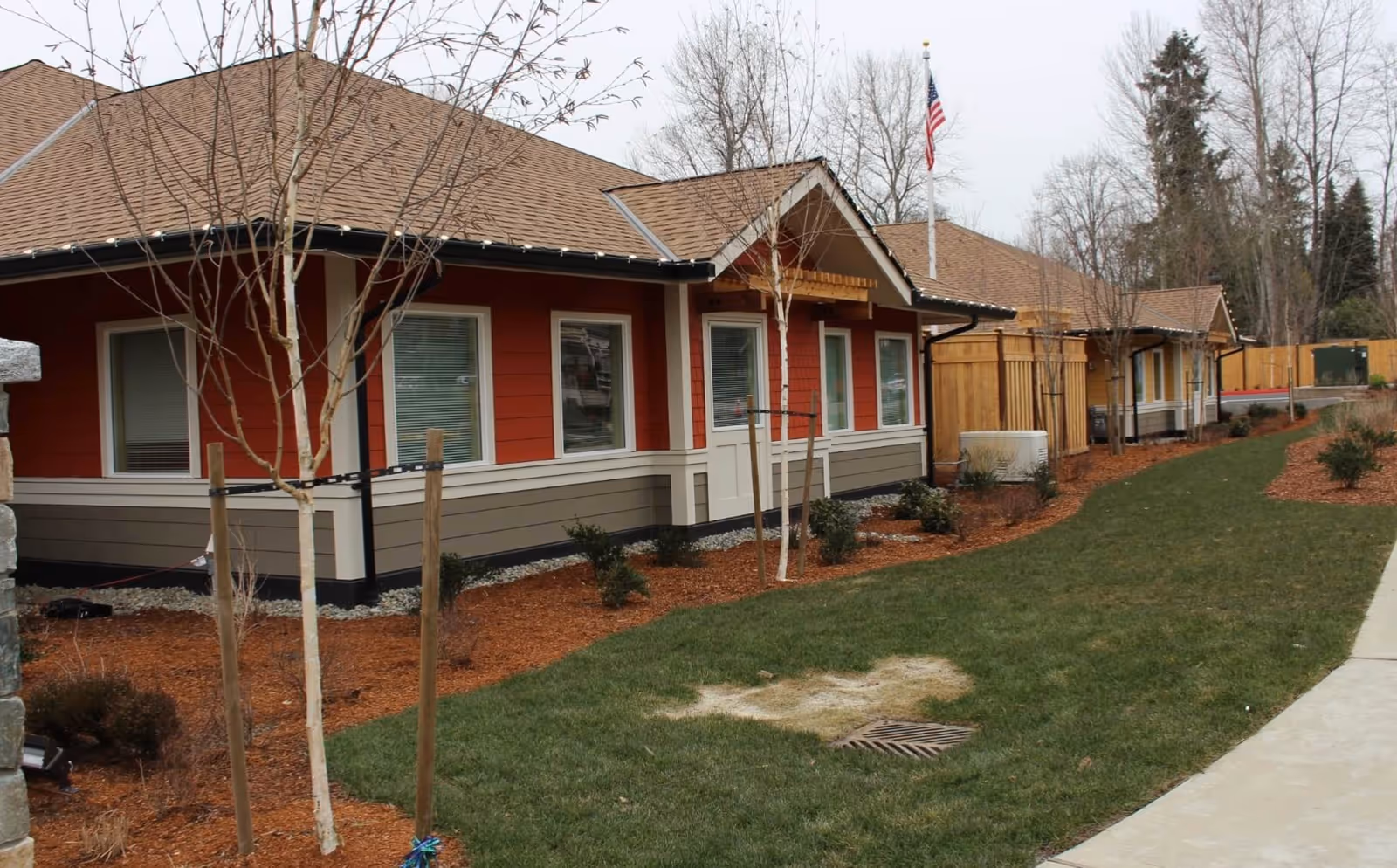 Exterior view of a single-story residential building with red and beige siding, multiple windows, and a brown shingled roof. There are young trees planted in mulch beds along the side of the building, a green lawn with a patch of dry grass, and a concrete walkway. An American flag is visible on a flagpole near the building.