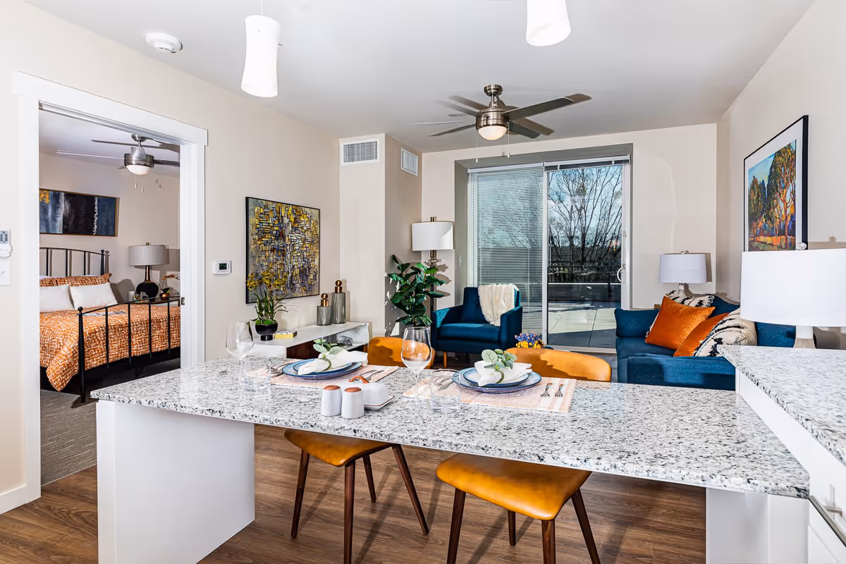 Open-plan apartment interior with a granite kitchen island set for dining, a living area with blue sofas and sliding glass door, and a bedroom visible through a doorway.