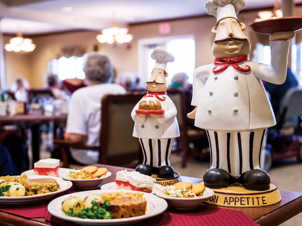 A dining room scene in a senior living facility with plates of food on a table in the foreground, including meatloaf, mashed potatoes, salad, crackers, and dessert. Two decorative chef statues are also on the table, one holding a loaf of bread and the other holding a covered dish. In the background, elderly people are seated and dining in a softly lit room.