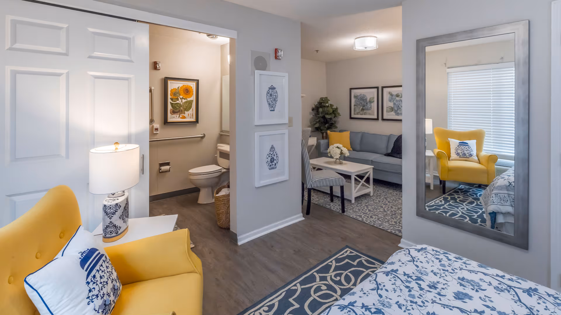 Studio-style senior living interior with a bed, yellow armchair, seating area, and an open bathroom.