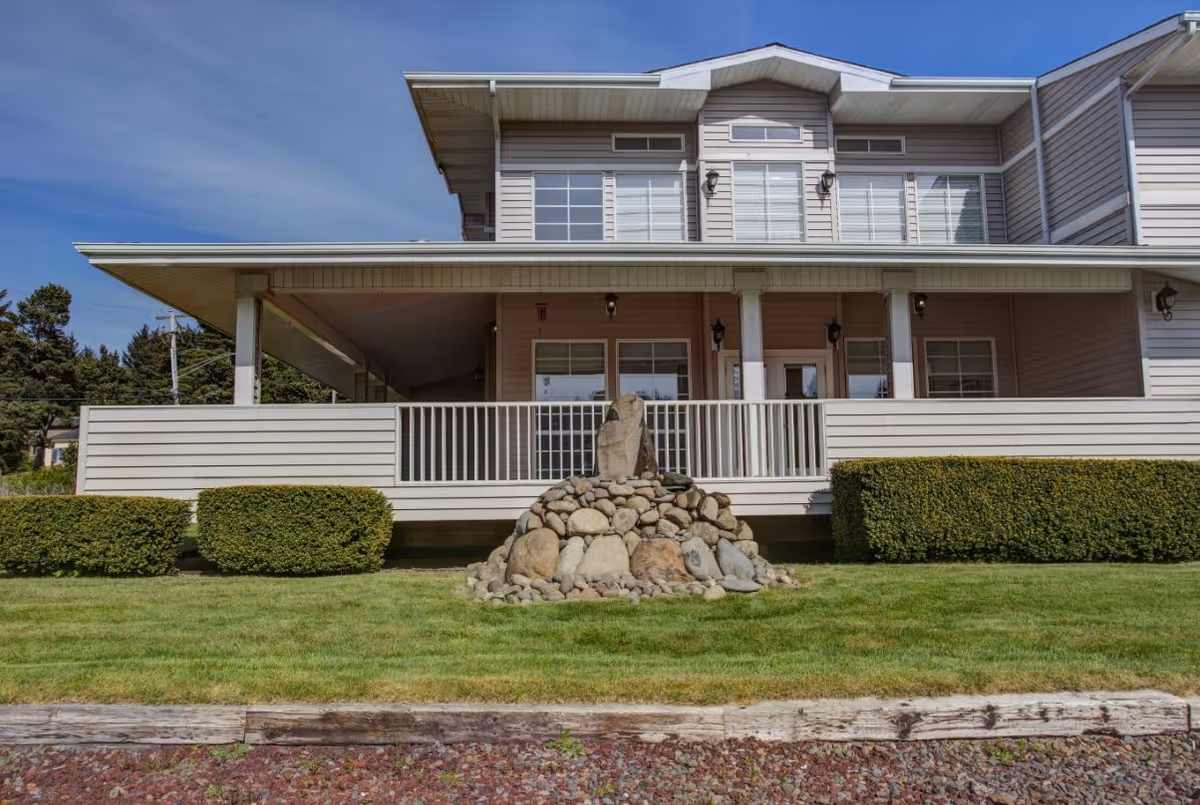 Exterior view of a two-story building with beige siding and white trim, featuring a covered porch with white railings. In front of the porch is a landscaped area with green grass, trimmed bushes, and a decorative rock arrangement. The sky is clear and blue.