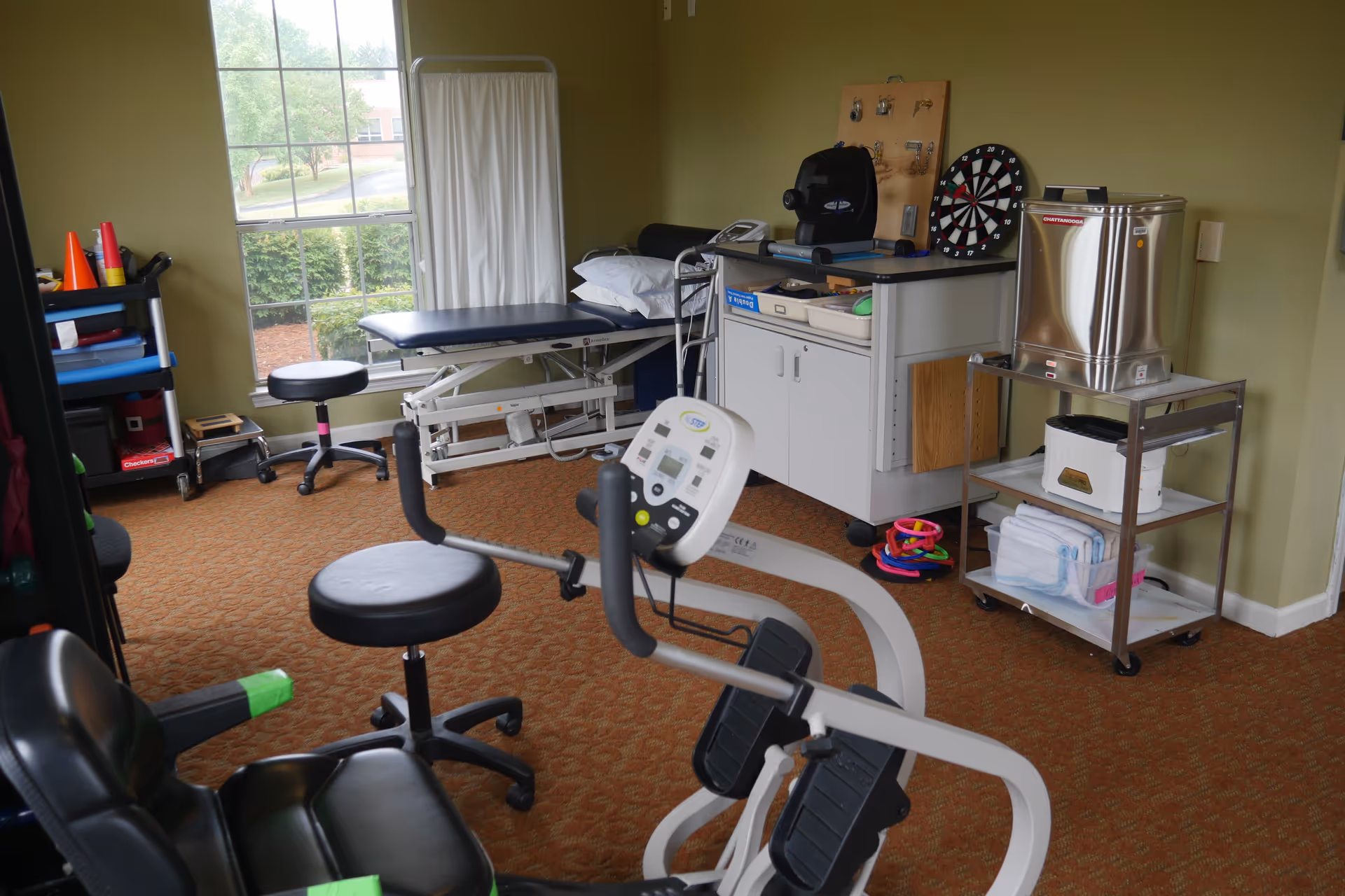 A therapy or rehabilitation room with exercise equipment, a treatment table with pillows, a stool, a dartboard on the wall, and various medical and therapy supplies on shelves and carts. A large window lets in natural light and shows greenery outside.