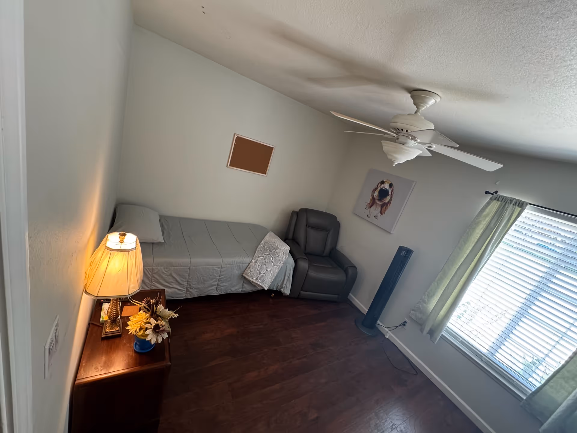 A small bedroom with a single bed covered in a gray bedspread, a wooden nightstand with a lamp and a flower vase, a gray armchair, a standing fan, a ceiling fan, a window with green curtains, and a wall decoration featuring a dog.