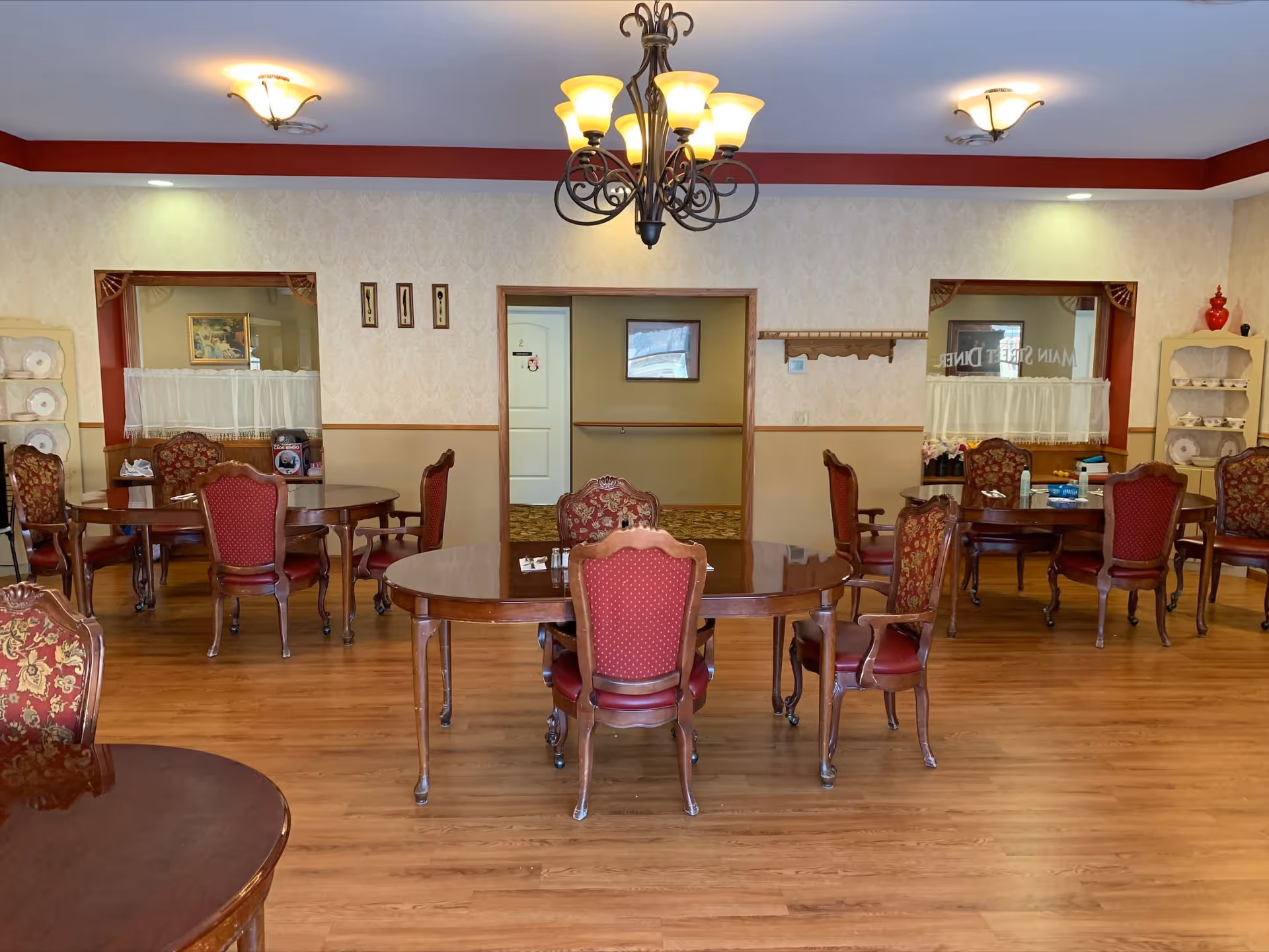 A dining room with multiple wooden tables and red upholstered chairs arranged neatly on a wooden floor. The walls have a beige patterned wallpaper with a red trim near the ceiling. There are two windows with white curtains and shelves displaying plates and teacups. A chandelier with multiple lights hangs from the ceiling, and there are additional ceiling lights. The room has a warm and inviting atmosphere.