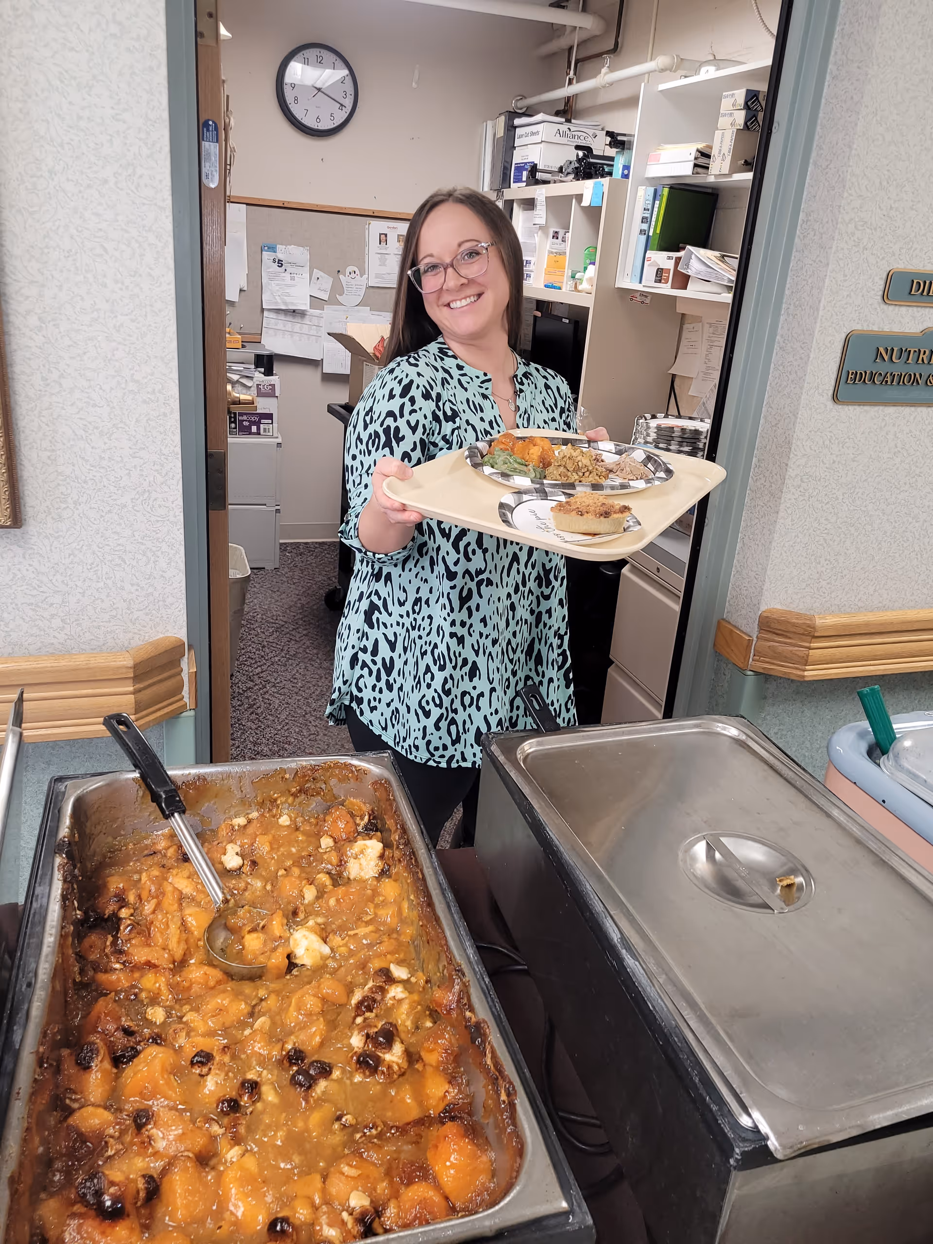 A smiling woman wearing glasses and a patterned blouse is holding a tray with a plate of food, standing in a doorway behind a buffet station with trays of food in a room that appears to be part of a healthcare facility.