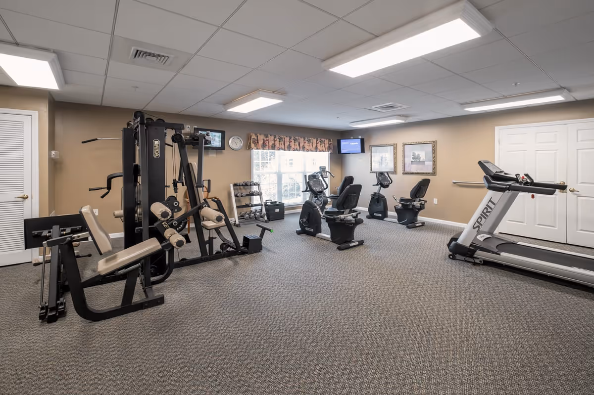 A fitness room with various exercise equipment including a multi-station weight machine, recumbent exercise bikes, a treadmill, and a rack of dumbbells. The room has beige walls, carpeted floor, ceiling lights, and two framed pictures on the wall. A window with a floral valance lets in natural light.