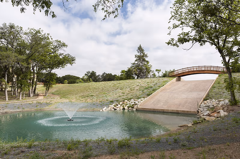 A small pond with a water fountain in the center, surrounded by grass and trees. A sloped concrete spillway leads up to a wooden footbridge crossing over the top. The sky is partly cloudy.