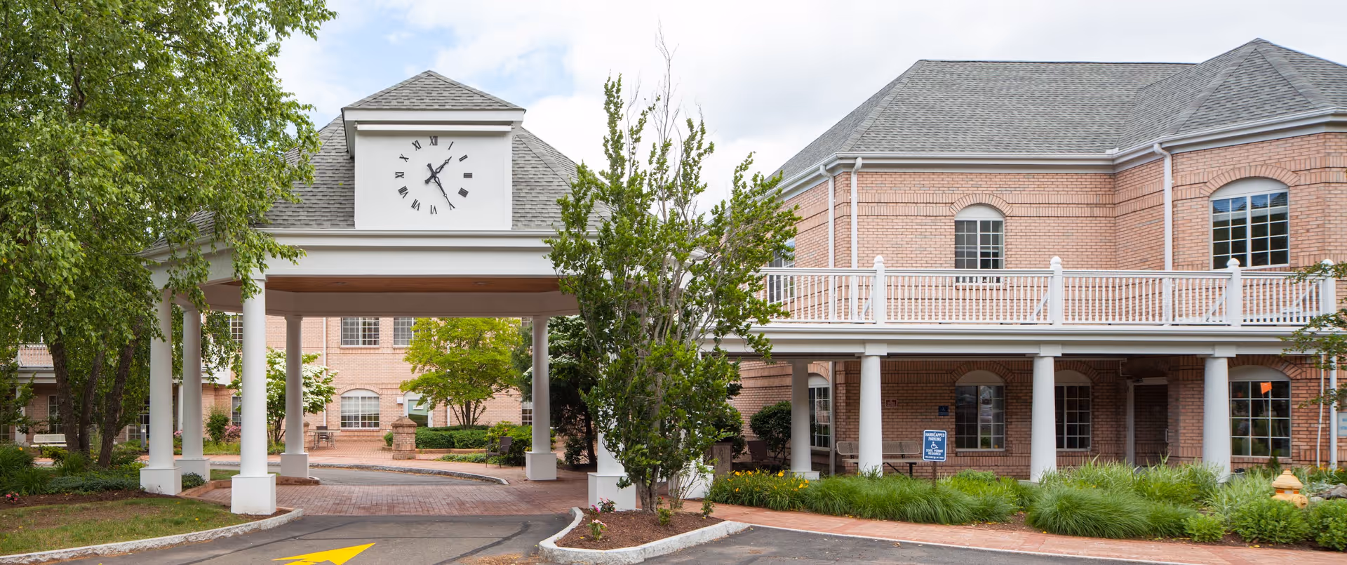 Exterior view of a senior living facility with a covered entrance featuring a clock tower. The building is made of light-colored brick with white railings and columns. There are trees and landscaped greenery around the entrance and parking area.