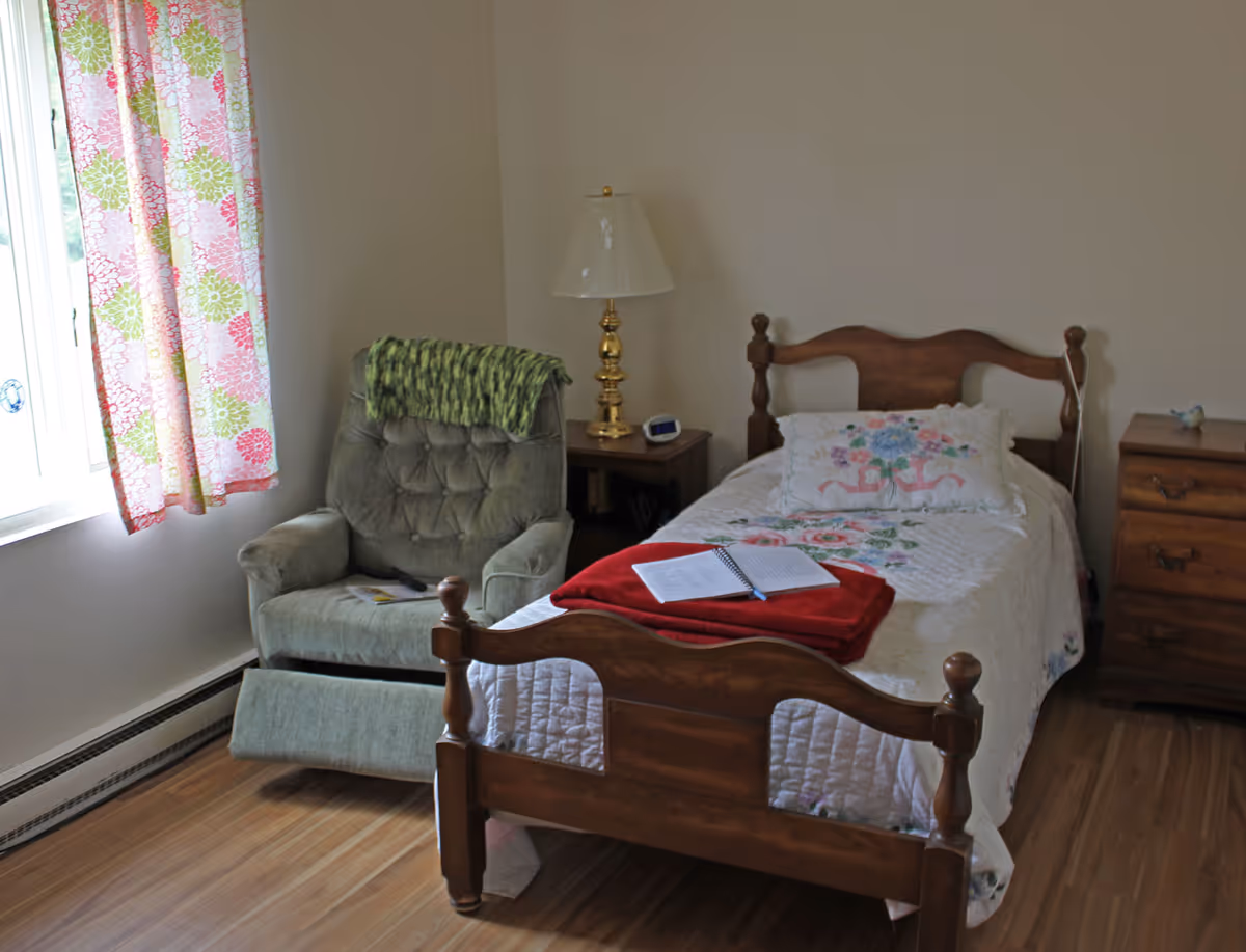 A cozy bedroom with a wooden bed featuring a floral quilt and matching pillow. A red blanket and an open notebook with a pen rest on the bed. Next to the bed is a wooden nightstand with a brass lamp and a digital clock. A green upholstered recliner chair with a green knitted throw is positioned near a window with floral curtains. The room has wooden flooring and light-colored walls.