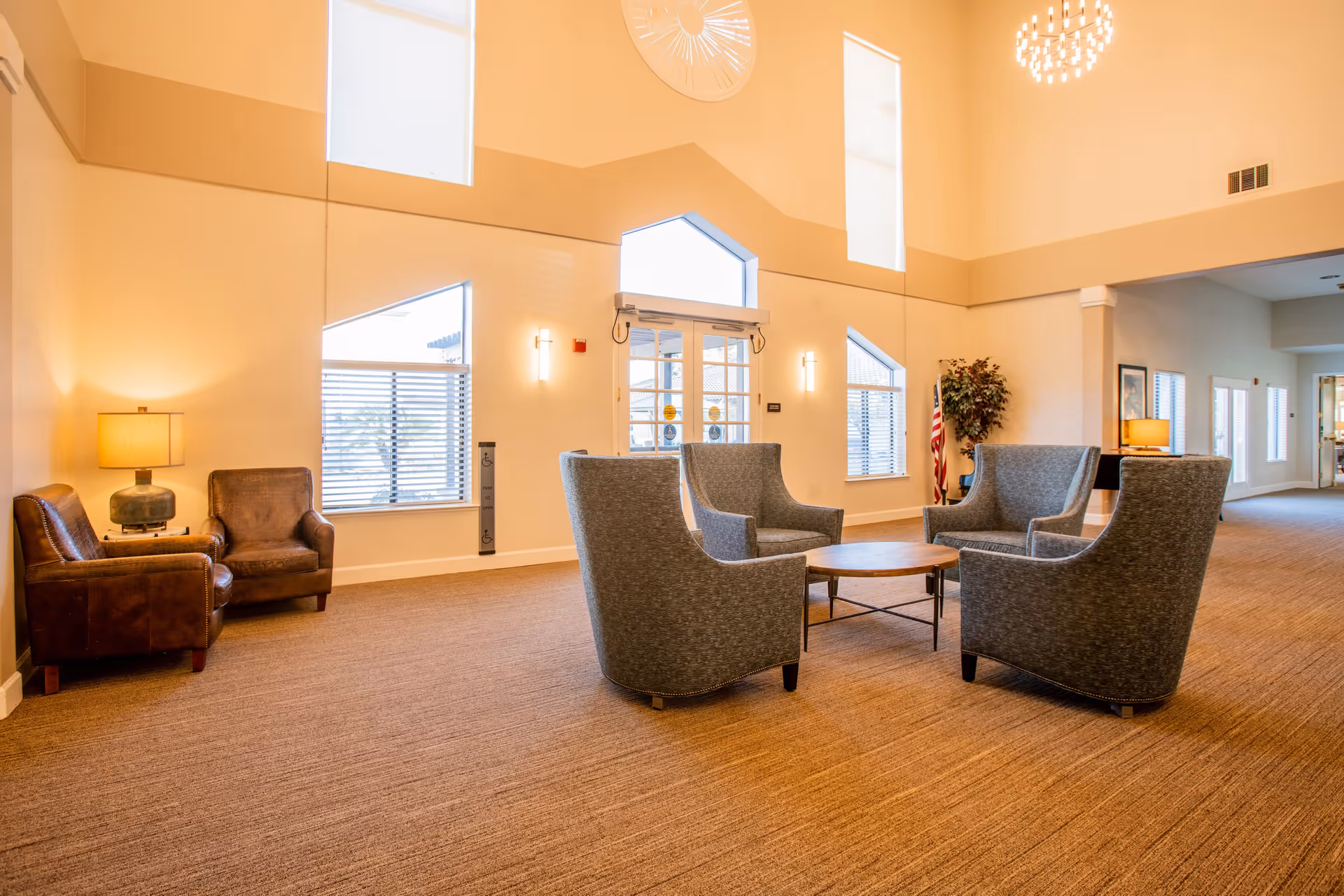 A spacious and well-lit common area with high ceilings featuring a circular chandelier. The room has a seating arrangement with four gray upholstered armchairs around a round wooden coffee table in the center. To the left, there are two brown leather armchairs with a lamp on a side table between them. Large windows with blinds allow natural light to enter, and an American flag is visible near a potted plant in the background. The walls are painted a warm beige color, and the carpet is a matching neutral tone.