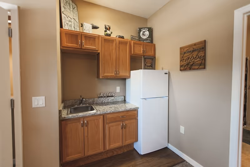 A small kitchen area with wooden cabinets, a granite countertop with a sink, and a white refrigerator. The walls are beige and decorated with small signs and artwork. The floor is wooden, and there is a light switch on the left wall.