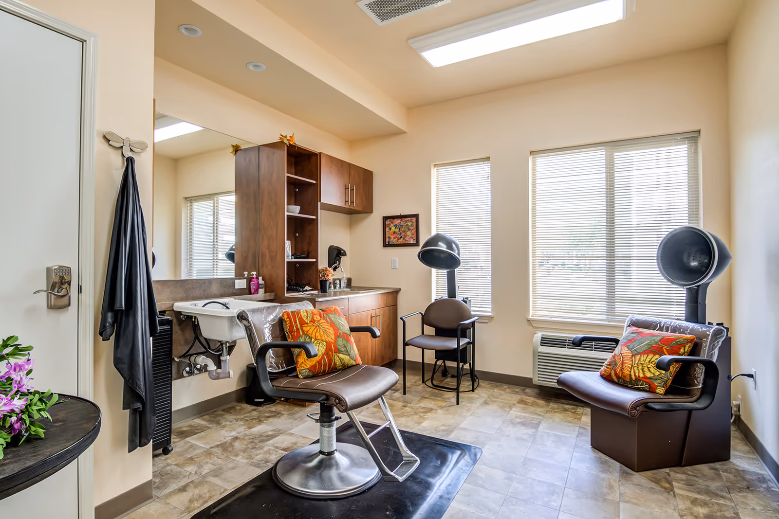 A bright salon-style room with styling chairs, hooded hair dryers, a wash basin, mirror and cabinetry.