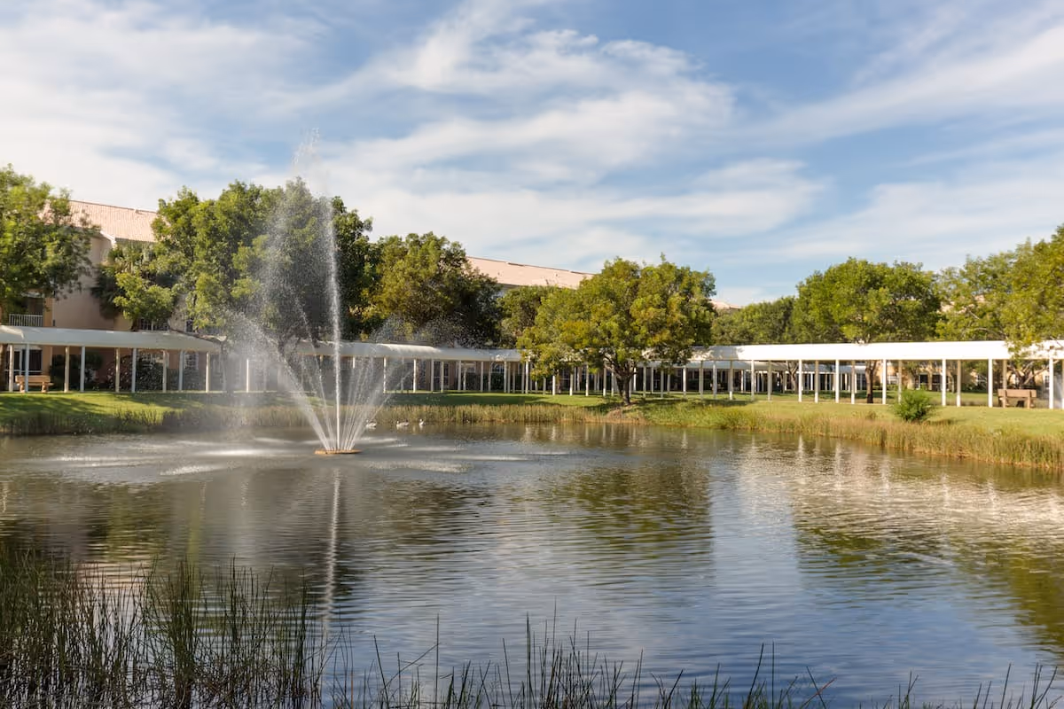 A serene outdoor scene at Aston Gardens At Pelican Marsh featuring a large pond with a central water fountain spraying water upwards. Surrounding the pond are green trees and a covered walkway with white pillars. The sky is partly cloudy with blue patches.