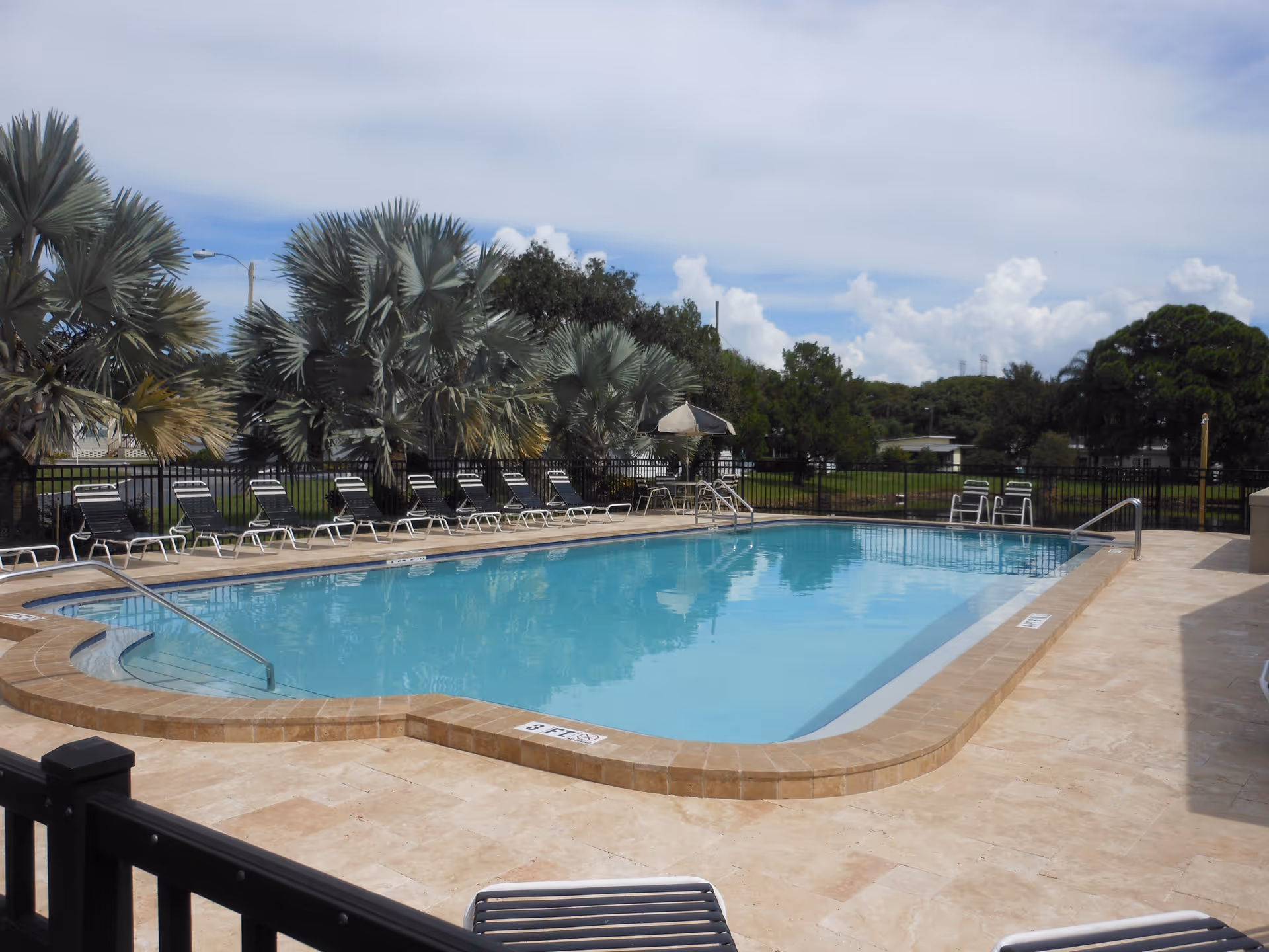 Outdoor swimming pool with clear blue water surrounded by a tiled deck. Several black and white lounge chairs are lined up along the poolside. There are palm trees and other greenery in the background under a partly cloudy sky.