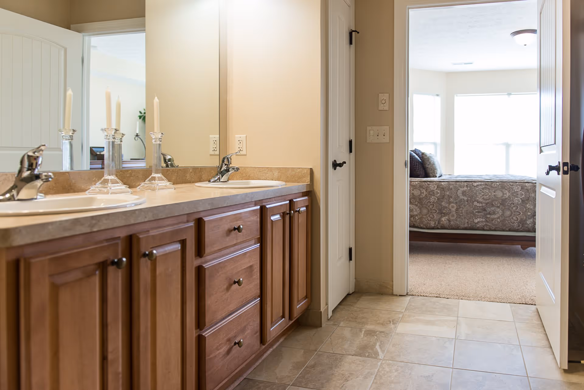 View of a bathroom with a double sink vanity featuring wooden cabinets and a large mirror. Two glass candle holders with white candles are placed on the countertop. The bathroom floor is tiled, and an open door leads to a bedroom with a bed covered in a patterned bedspread and large windows letting in natural light.