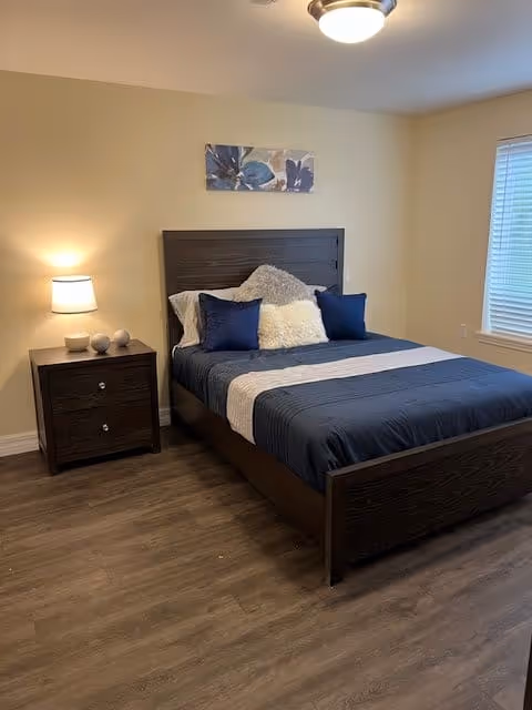 A bedroom with a wooden bed frame and a neatly made bed featuring dark blue bedding, white and blue pillows, and a fluffy white accent pillow. There is a wooden nightstand with a lamp and decorative items on it to the left of the bed. A window with blinds is on the right wall, and a floral artwork hangs above the bed. The room has wood flooring and beige walls.