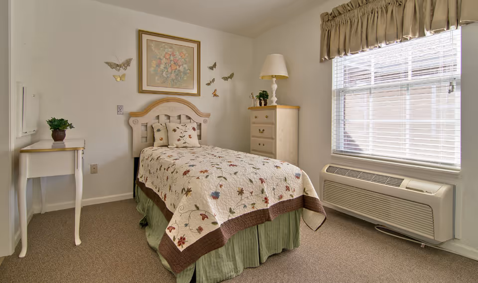 A small, cozy bedroom with a single bed covered in a floral quilt and matching pillows. The room features a wooden headboard, a small white side table with a potted plant, a wooden dresser with a lamp and decorative items, a window with blinds and a beige valance, and beige carpeted flooring. Butterfly wall decorations and a framed floral painting hang above the bed.