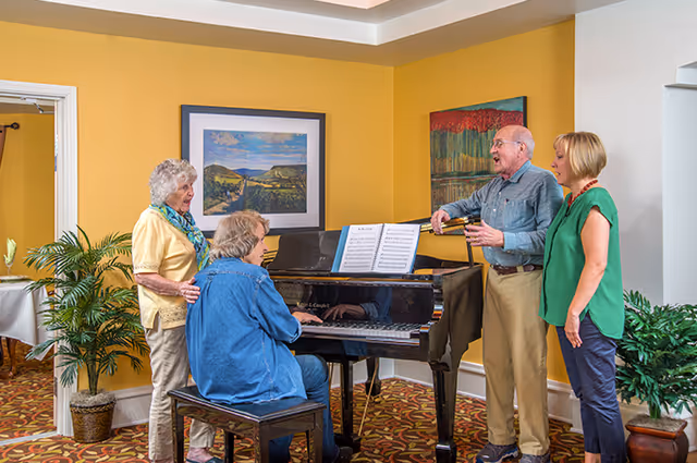 Four seniors gathered around a black grand piano in a bright common room, with one person playing and three standing and singing.