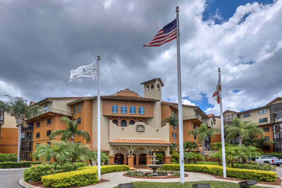 Entrance of a multi-story Mediterranean-style senior living building with flagpoles, palm trees, and a central fountain.