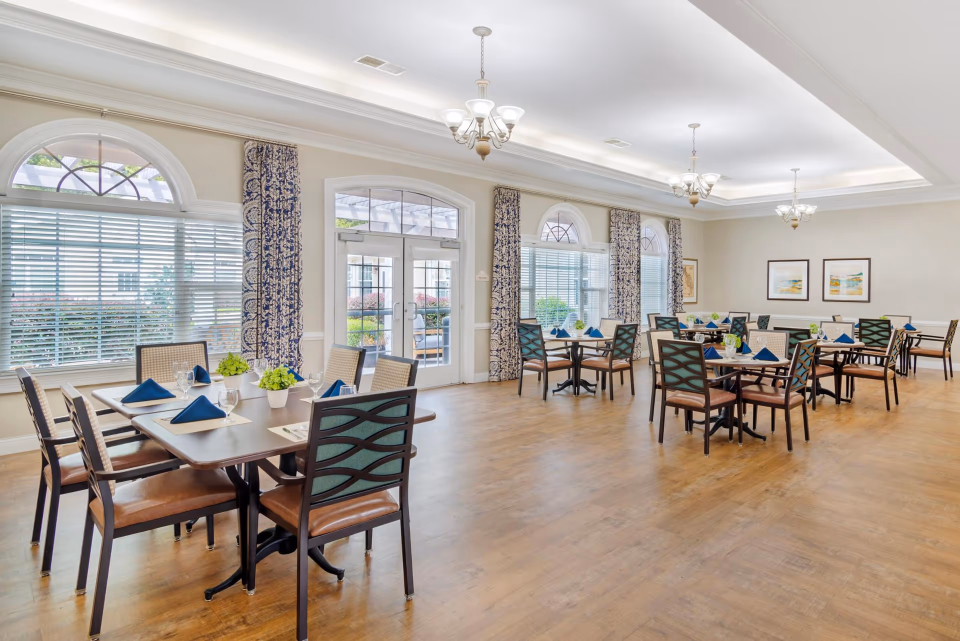 Spacious, well-lit dining room with multiple tables and chairs set with blue napkins, large arched windows, and glass doors.