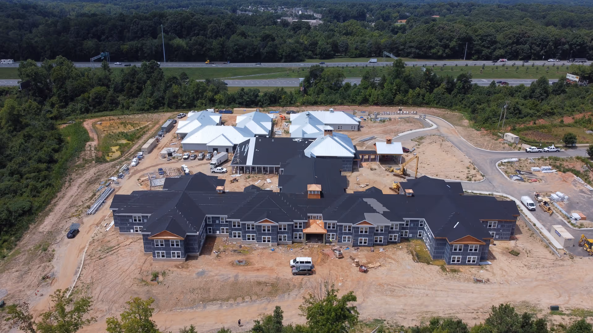 Aerial view of Arbor Ridge at Huntersville senior living facility under construction, showing multiple connected buildings with black and white roofs, surrounded by dirt and construction equipment, with a highway and forested area in the background.