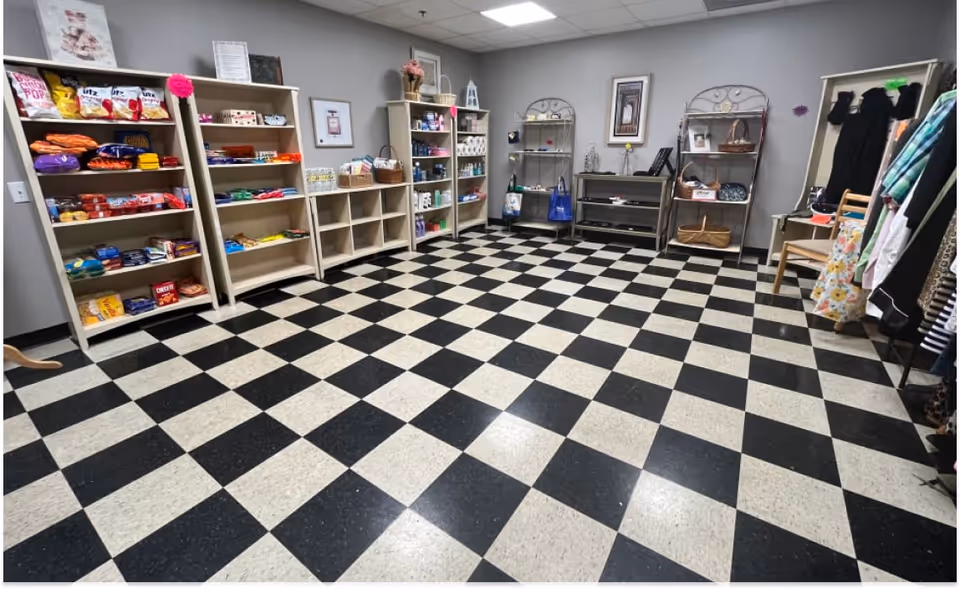 Interior of a small shop or boutique with black-and-white checkered tile floor and shelves stocked with snacks, clothing, and household items.