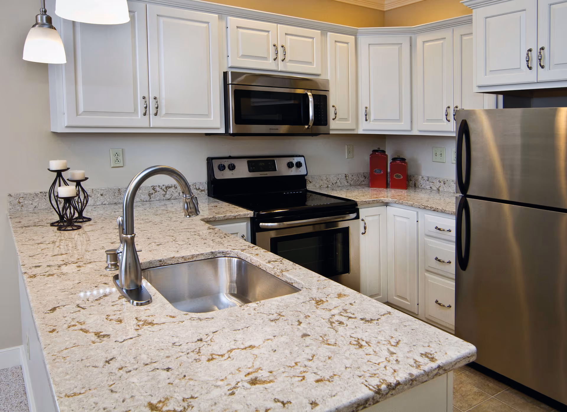 Modern kitchen with white cabinets, granite countertops, stainless steel sink with a curved faucet, stainless steel refrigerator, stove, and microwave. There are two red canisters on the countertop and a decorative candle holder with three candles.