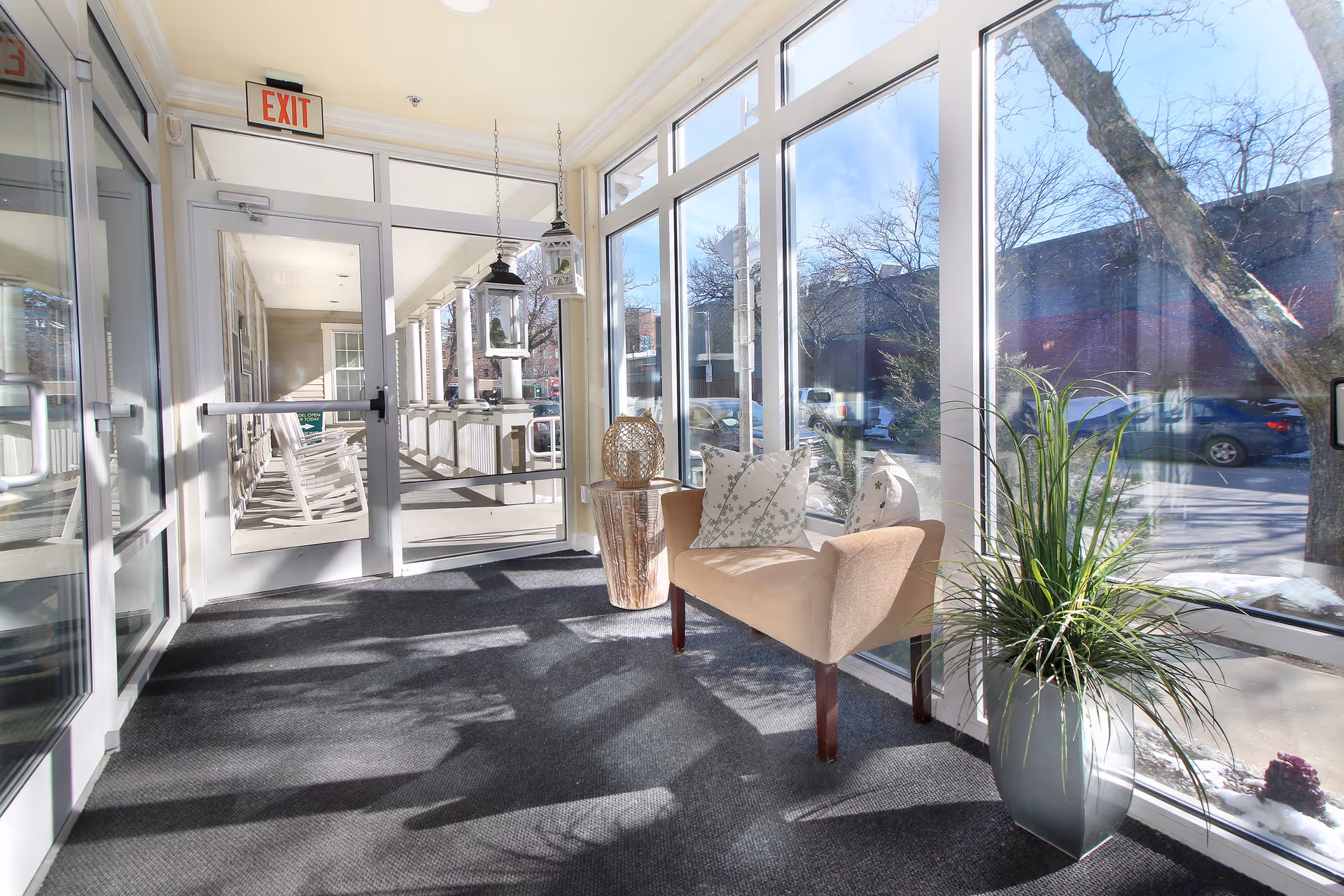 A bright sunlit indoor seating area with large floor-to-ceiling windows showing an outdoor view with trees and parked cars. The area features a beige upholstered bench with two patterned pillows, a small wooden side table with a decorative lantern, a tall potted plant, and hanging lanterns near a glass exit door leading to a porch with white rocking chairs.