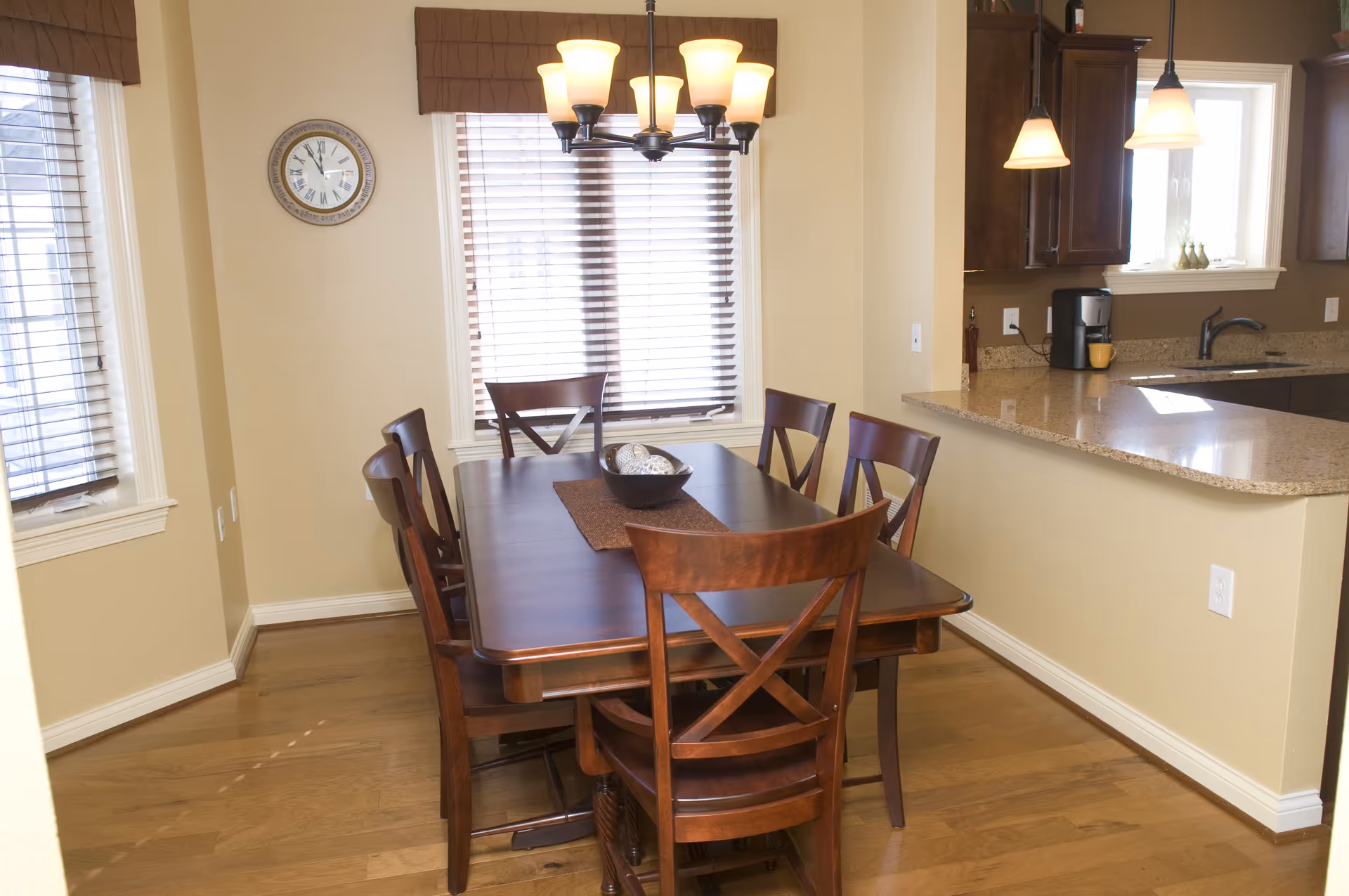 Dining area with a wooden table and six chairs beside an open kitchen with a granite countertop and pendant lights.