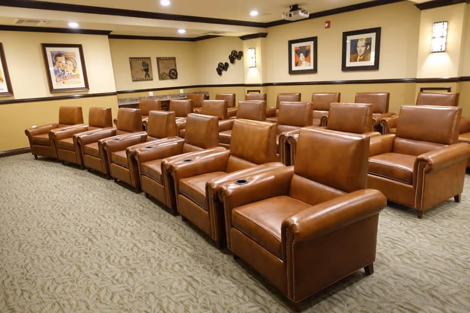 A room with multiple rows of brown leather armchairs arranged theater-style facing a screen or stage. The walls are decorated with framed vintage movie posters and film reels, and the room is well-lit with ceiling lights and wall sconces.