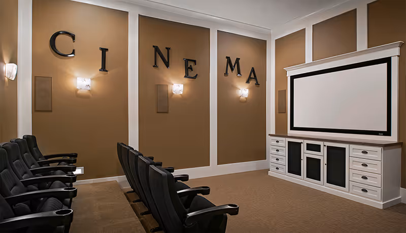 A small cinema room with two rows of black theater seats facing a large white projection screen mounted on a white cabinet with drawers. The walls are painted brown with white trim, and large black letters spelling 'CINEMA' are mounted on the wall above the seats. Soft wall sconces provide lighting.