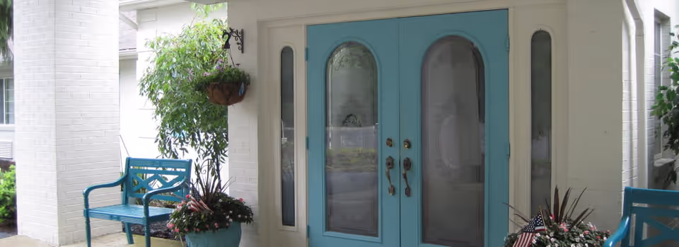 Entrance area of a facility with double blue doors featuring glass panels, flanked by white brick walls. There are blue benches on either side of the entrance and potted plants with flowers and small American flags near the doors. A hanging plant is visible on the left side.