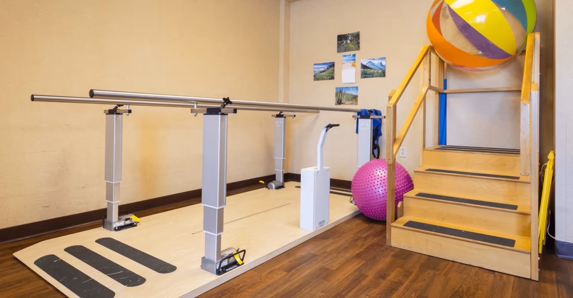 A physical therapy or rehabilitation room with parallel bars for walking exercises, a set of wooden stairs with handrails, a large colorful beach ball, and a pink spiked exercise ball. The walls have several nature-themed pictures and a calendar.